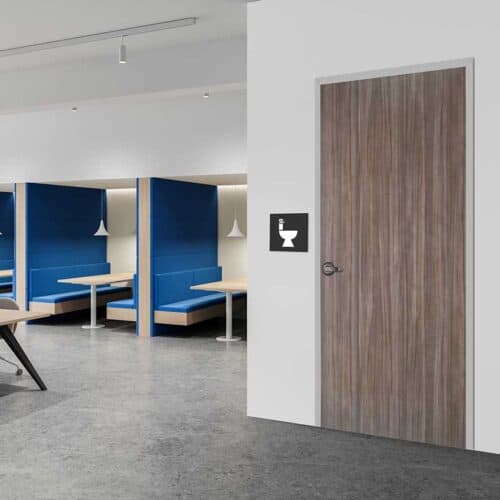 A closed wooden restroom door with a toilet sign, set in a FlexFit™ Field-Adjustable Interior Aluminum Door Frame, sits next to blue booth seating in a modern office or public space.