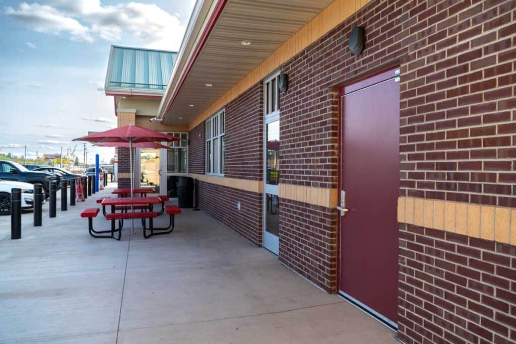 Outdoor seating area with red picnic tables and umbrellas beside a brick building featuring a maroon national account commercial door solution on a clear day.