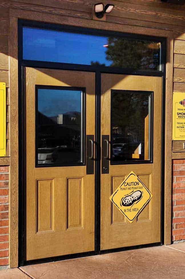 Two wooden doors with glass panels; a yellow caution sign with a peanut icon warns of peanut dust in the area—ideal for businesses seeking national account commercial door solutions.