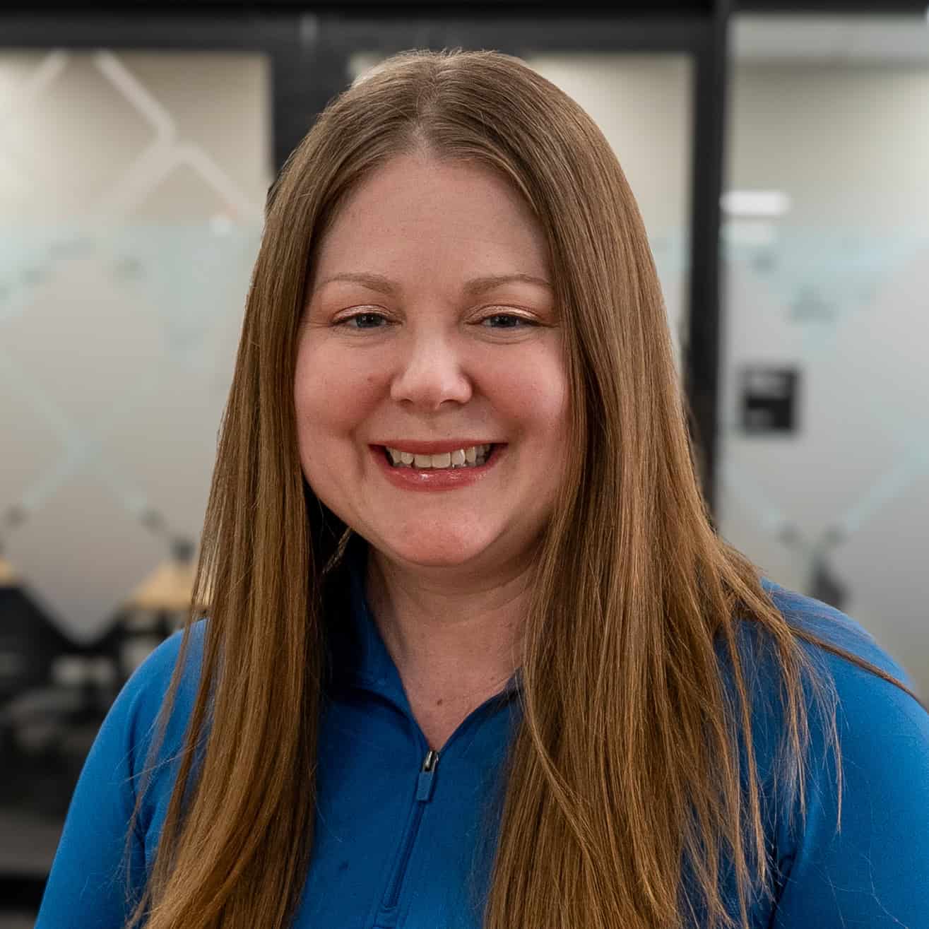 Woman with long straight light brown hair, wearing a blue zip-up top, smiling in an office setting with frosted glass panels in the background, showcasing the friendly spirit of our team.