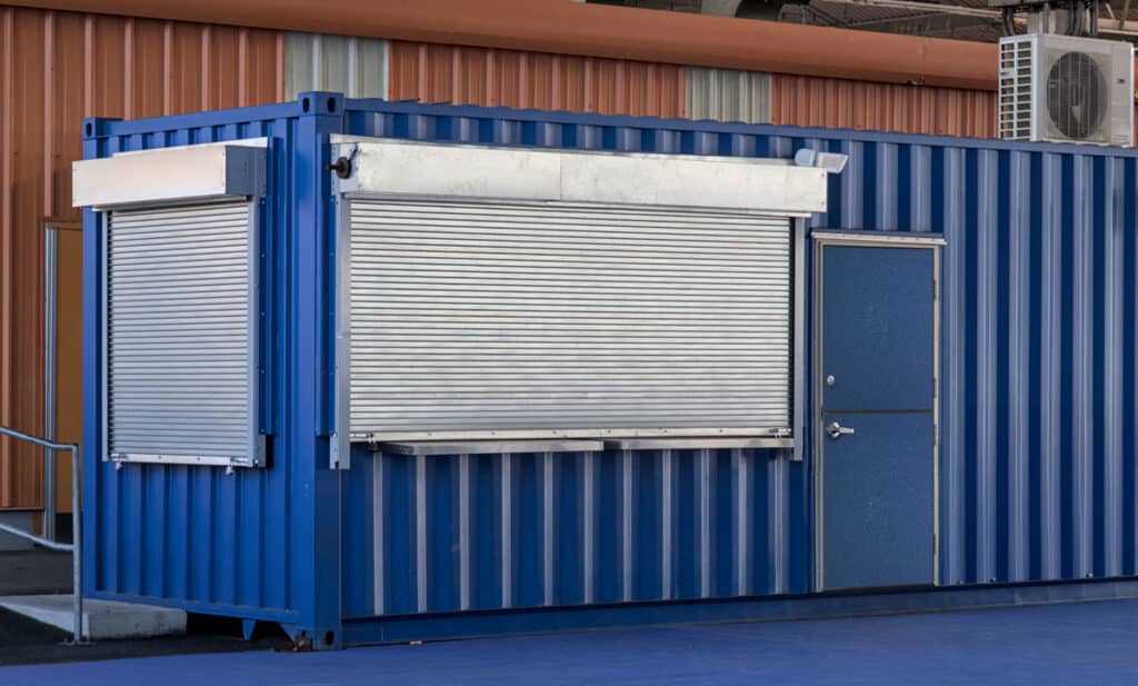 A blue shipping container with closed metal shutters and a national account commercial door, set on a blue-painted ground, featuring an air conditioning unit mounted on top.