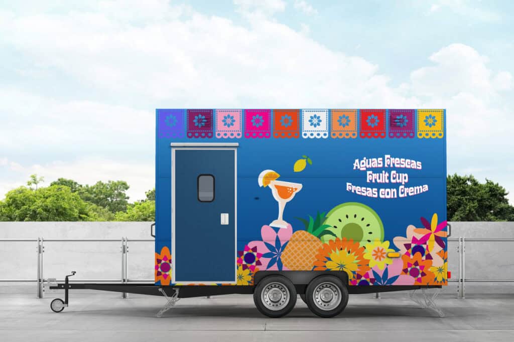 A colorful food trailer with fruit and floral designs advertises "Aguas Frescas, Fruit Cup, Fresas con Crema," showcasing the versatility of OEM Account Solutions for Modular and Prefabricated Mobile Facilities against a clear sky and tree background.