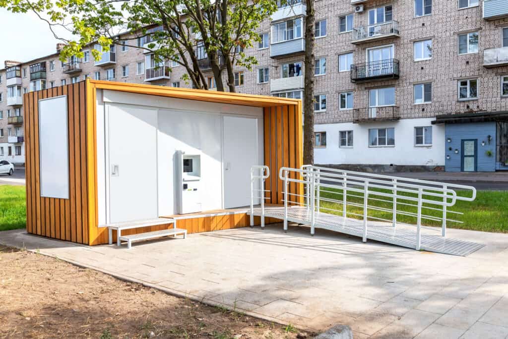 Small, modern kiosk with a wooden exterior, white doors featuring national account commercial door solutions, and an accessibility ramp, located outside a multi-story apartment building.