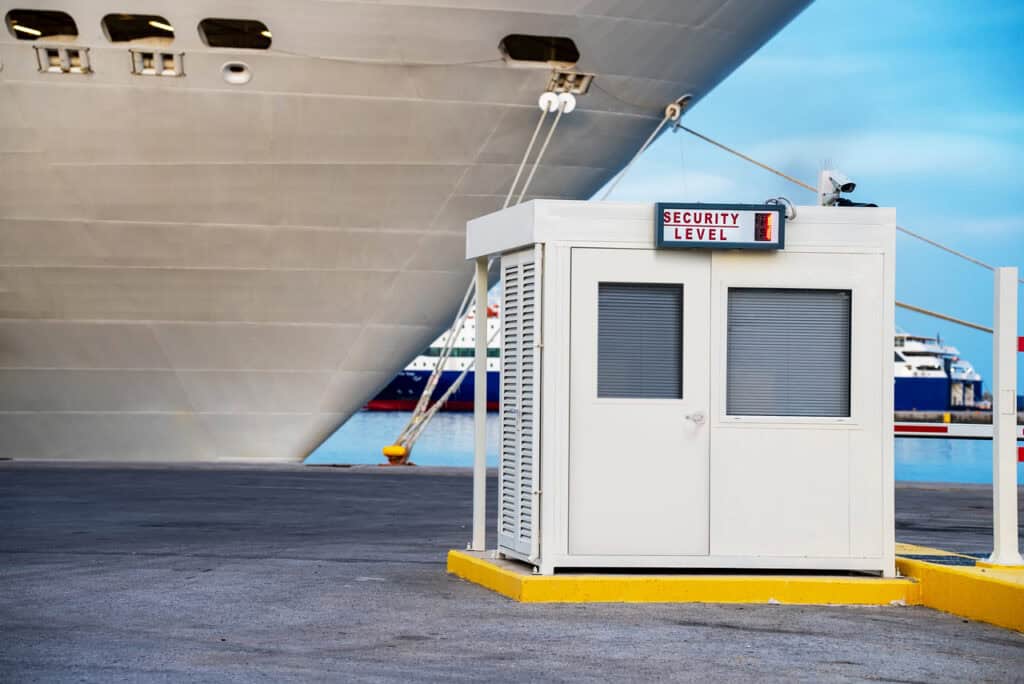 A small white security booth with a "Security Level 1" sign, designed using OEM Account Solutions for Modular and Prefabricated Mobile Facilities, is positioned near a docked cruise ship in a harbor.
