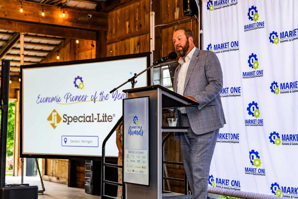 A man in a gray suit speaks at a podium during an awards event. A screen behind him displays “Economic Pioneer of the Year, Special-Lite, Decatur, Michigan,” celebrating Special-Lite’s achievements in the Market One sector.