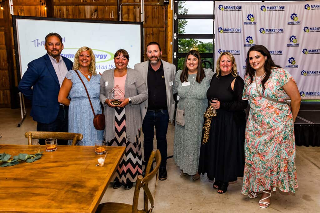 Seven people stand indoors posing for a group photo; one person in the center holds an Economic Pioneer Award. A "Market One" backdrop and a presentation screen are visible in the background, highlighting Special-Lite’s achievements.