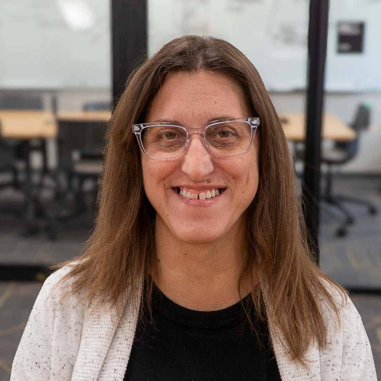 A woman with long brown hair and clear glasses is smiling while standing in an office with glass walls and empty chairs in the background, reflecting the welcoming atmosphere of our team.