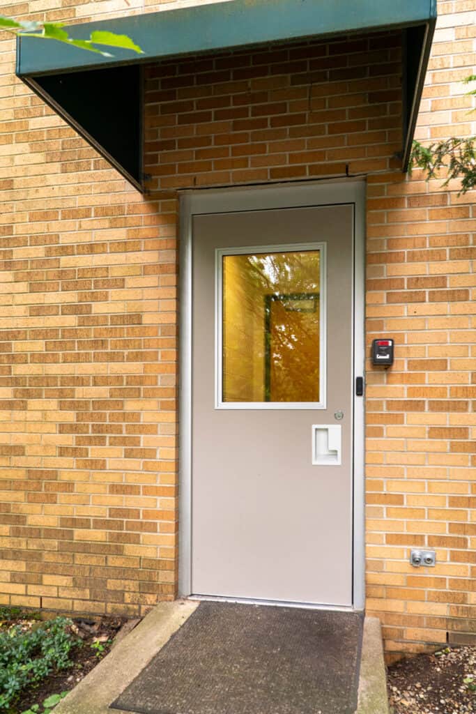 A gray door with a window and card reader—highlighting enhanced security and modern entrance systems—is set in a brick wall under a green awning, with a black doormat on the concrete step.